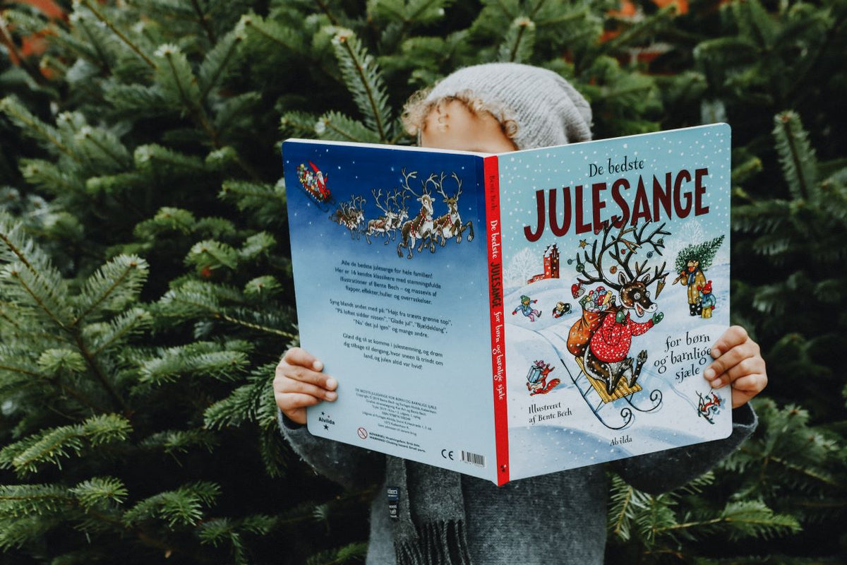 Barn med hue læser julebog "De Bedste Julesange" foran grønne juletræer.