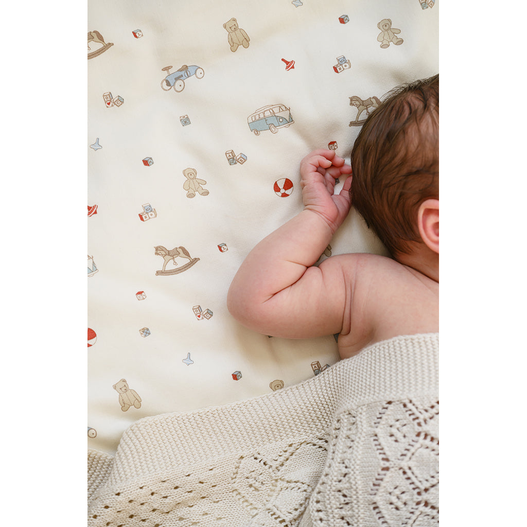 Babyhoved og arm på økologisk babysengetøj med legetøjsprint og beige tæppe